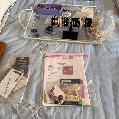 Collection of crafting supplies, crochet patterns, knit blankets, and costume jewelry displayed on a bed, including a white serving tray...