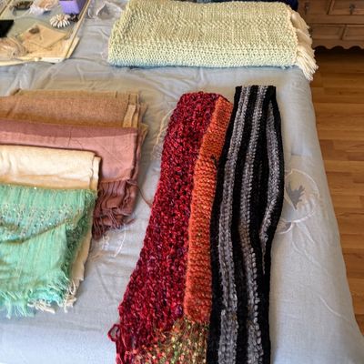 Collection of assorted winter textiles, including handmade crocheted blankets, knit scarves, and folded pashmina-style wraps displayed on...