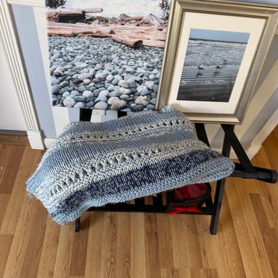 Hand-knitted blue and grey striped throw blanket displayed on a black luggage rack alongside two coastal-themed framed art pieces.