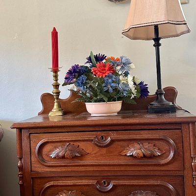 Sale Photo Thumbnail #119: Antique wooden three-drawer chest with carved leaf pulls, displayed alongside tabletop decor, a table lamp, and wall-mounted decorative plates.