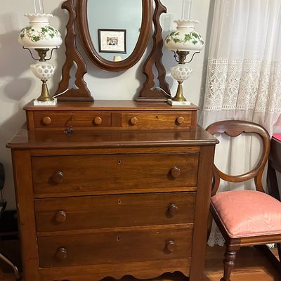 Sale Photo Thumbnail #87: Antique wooden dresser with an attached oval mirror, flanked by a pair of brass and hand-painted milk glass hurricane lamps, with an adjacent Victorian balloon-back chair.