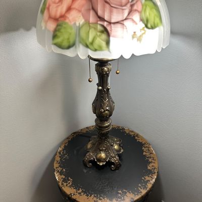 Sale Photo Thumbnail #25: Vintage table lamp featuring an ornate cast metal base and a scalloped glass shade with hand-painted pink rose motifs, resting on a distressed round accent table.