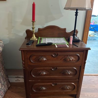 Sale Photo Thumbnail #20: Vintage carved wood three-drawer nightstand or small chest featuring leaf motif details, an arched crest rail, and a table lamp on top.