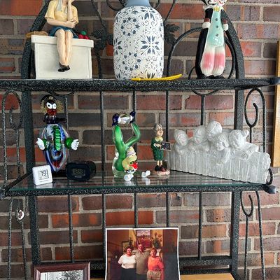 Sale Photo Thumbnail #2: Black wrought iron baker's rack displayed with assorted collectibles, Murano-style glass clown figurines, ceramics, decorative vases, and framed photographs.