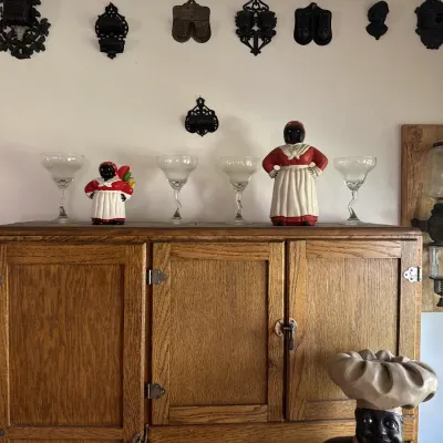 Sale Photo Thumbnail #37: Vintage oak icebox or kitchen cabinet topped with Black Americana collectibles, margarita glasses, and cast-iron wall hangings.