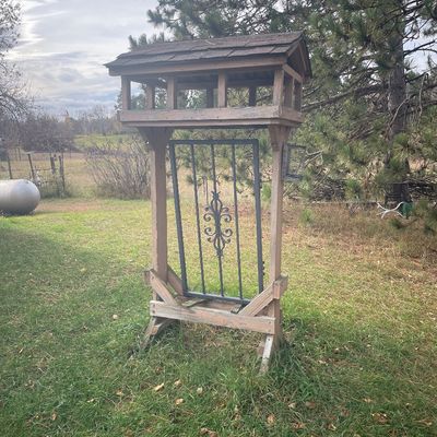 Sale Photo Thumbnail #178: Rustic wooden covered bird feeder stand featuring a shingled roof and a wrought iron scrollwork accent panel.