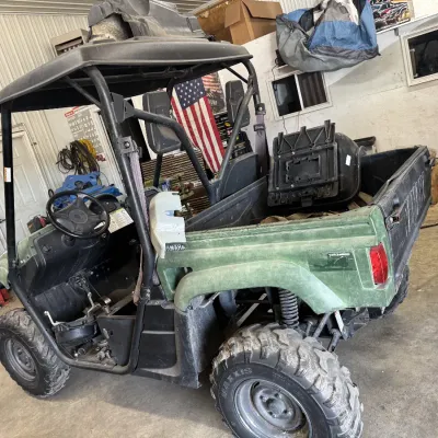 Sale Photo Thumbnail #100: Yamaha Rhino side-by-side utility vehicle in green, showing visible wear and missing body panels, equipped with a roof canopy and knobby Maxxis tires.