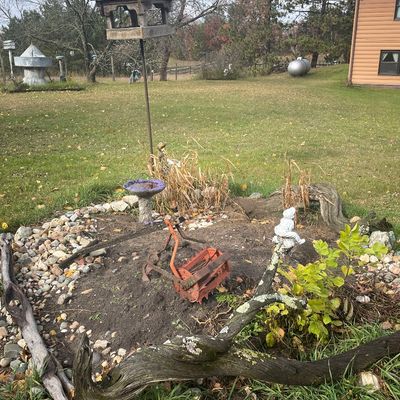 Sale Photo Thumbnail #168: Rustic garden landscape featuring a wooden birdhouse on a pole, a vintage orange push reel lawnmower, small garden statues, and a birdbath surrounded by river rocks and drift logs.
