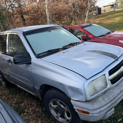 Sale Photo Thumbnail #153: Silver Chevrolet Tracker SUV showing exterior wear and minor body damage, parked outdoors beside a red sedan.