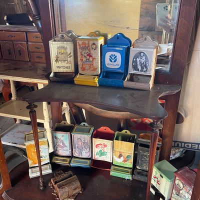 Sale Photo Thumbnail #191: Collection of vintage tin match safes displayed on a dark wood two-tier vanity shelf with a mirror.