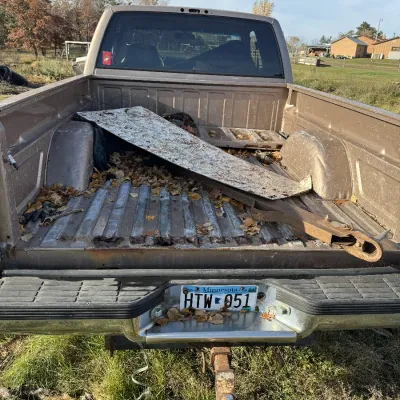 Sale Photo Thumbnail #127: Open bed of a tan pickup truck showing rust, fallen leaves, and miscellaneous metal debris, fitted with a Minnesota license plate and tow hitch.