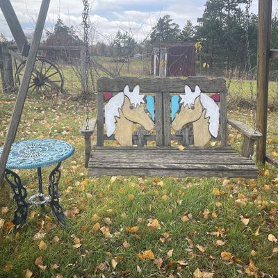 Sale Photo Thumbnail #173: Rustic wooden porch swing featuring painted horse cutouts on the backrest, suspended on a wooden A-frame alongside a small light blue cast iron side table.
