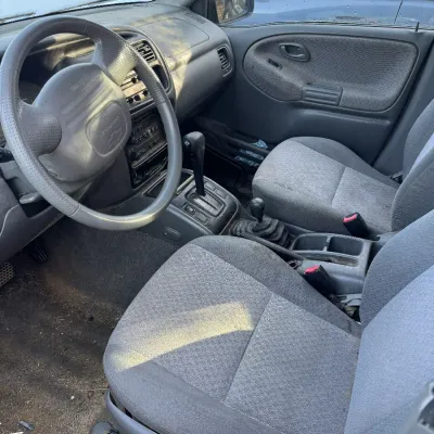 Sale Photo Thumbnail #157: Interior view of a late-1990s or early-2000s Chevrolet Tracker, featuring grey cloth bucket seats, automatic transmission shifter, and four-wheel-drive gear selector.