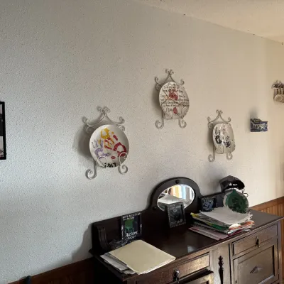 Sale Photo Thumbnail #45: Collection of decorative wall plates with wire hangers mounted above a dark wood sideboard mirror cabinet, accompanied by framed artwork and a corner display rack.