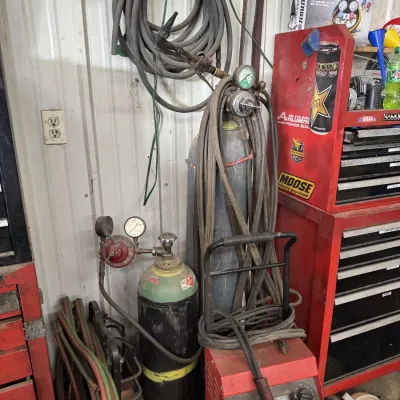 Sale Photo Thumbnail #70: Oxy-acetylene welding setup featuring compressed gas tanks with regulators, torch attachment, coiled hoses, and a red portable welding unit in a workshop setting.