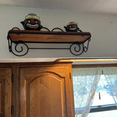 Sale Photo Thumbnail #34: Pair of vintage clown face novelty teapots with wire handles, displayed on a wall-mounted wrought iron and wood shelf.