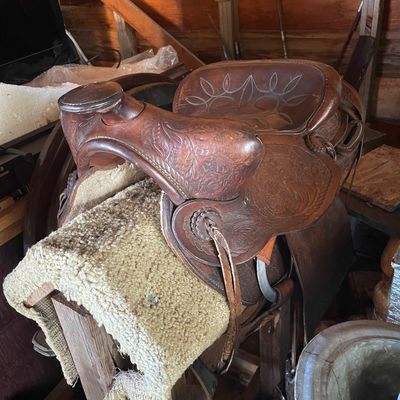 Sale Photo Thumbnail #196: Vintage floral-tooled leather Western saddle resting on a wooden stand with fleece padding, showing natural patina and wear.