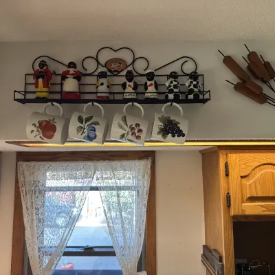Sale Photo Thumbnail #31: Wall-mounted wrought iron spice shelf featuring vintage Black Americana salt and pepper shakers, hanging ceramic fruit mugs, and a wooden cattail wall decor piece.