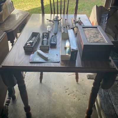 Sale Photo Thumbnail #201: Wooden drop-leaf dining table arranged with a collection of pocket knives, fixed-blade hunting knives, daggers, and a upholstered wooden box.