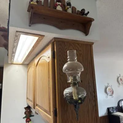 Sale Photo Thumbnail #28: Vintage wall-mounted clear glass oil lamp attached to a wooden cabinet, displayed alongside a decorative wooden shelf above.