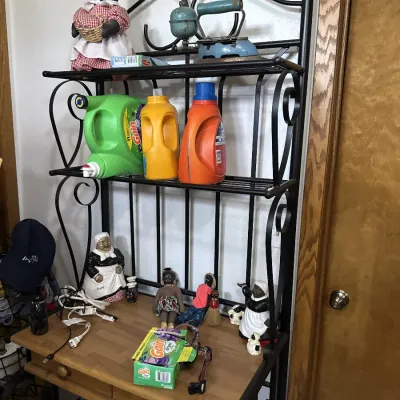 Sale Photo Thumbnail #20: Black wrought iron baker's rack with a wooden tabletop, displaying various laundry supplies, vintage iron collectibles, and Black Americana dolls and figurines.