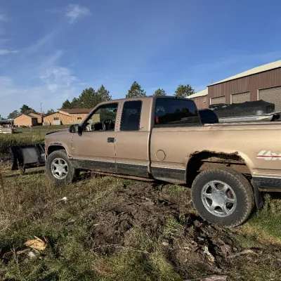 Sale Photo Thumbnail #128: Tan late-1990s Chevrolet or GMC extended cab 4x4 pickup truck fitted with a front snow plow attachment, showing visible body rust along the rear wheel well.
