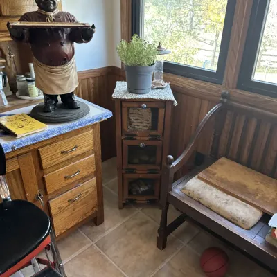 Sale Photo Thumbnail #25: Eclectic kitchen corner featuring a vintage wooden chef statue holding a tray, a oak cabinet with blue trim, a three-tier screen-door vegetable bin, and a wooden bench.