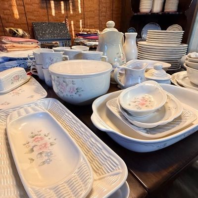 Vintage white ceramic dinnerware set featuring delicate pink floral motifs, serving platters, bowls, cups, and a butter dish arranged on...