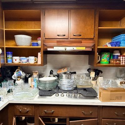 Assorted vintage kitchenware, including glassware, mugs, cookware, and small appliances, displayed on counters and open wooden cabinetry.
