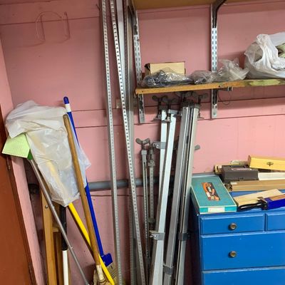 Assortment of long aluminum bar clamps leaned against a wall in a workshop area alongside cleaning tools and a blue chest of drawers.