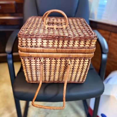 Vintage woven wicker sewing basket or picnic caddy featuring a top lid handle and a fold-down front handle, resting on a black chair.