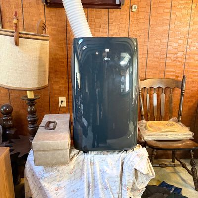Modern dark grey LG portable air conditioner unit with exhaust hose, shown alongside a vintage table lamp and wooden side chair.
