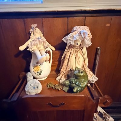 Vintage soft-sculpture rabbit dolls resting on a ceramic pitcher and wooden nightstand, alongside ceramic frog figurines and a plush cat.