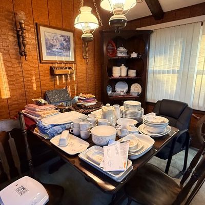 A wood dining table set surrounded by various home goods, including stacks of linens, porcelain dinnerware, serving dishes, and teacups,...