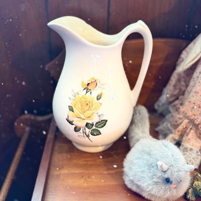 Vintage cream ceramic pitcher featuring a printed yellow rose floral motif, displayed alongside small decor items on a wooden surface.