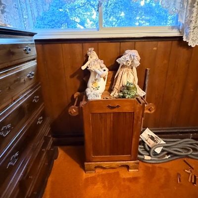 Vintage wooden dry sink stand holding decorative dolls, a ceramic pitcher, and a frog figurine, positioned next to a tall wooden dresser.