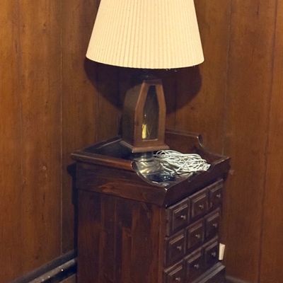 Vintage dark pine nightstand featuring multiple small drawers, paired with a lantern-style table lamp with a pleated fabric shade.