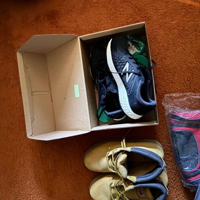 Pair of black New Balance sneakers in an open cardboard box alongside tan work boots resting on shag carpet.