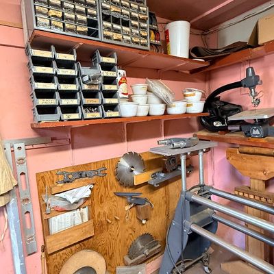 Workshop storage setup featuring metal drawer organizers, stacked part bins, table saw blades mounted on wall panels, and stationary...