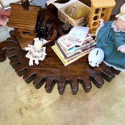 Rustic gear-shaped wooden side table holding a log cabin lamp, cookbooks, small pig figurines, and glassware.