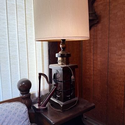 Vintage dark wood table lamp with brass accents and a tall drum shade, displayed alongside a decorative miniature wooden harp.