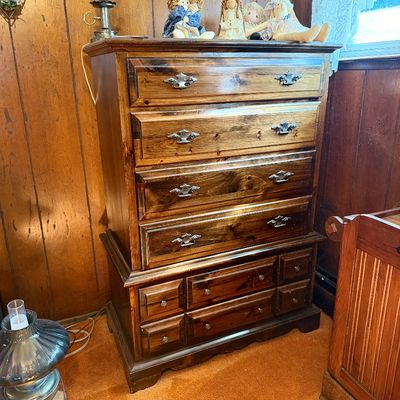 Vintage dark-stained pine chest-on-chest tall dresser with brass batwing hardware, topped with decorative dolls and an oil lamp.