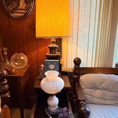 Vintage table lamp with a textured gold drum shade, paired with a white milk glass hurricane-style lamp on a dark wood end table next to...