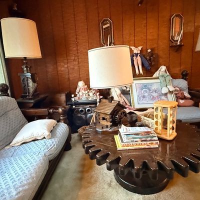 Vintage wood gear-shaped coffee table surrounded by a blue upholstered sofa, armchair, and table lamps in a paneled living room setting.