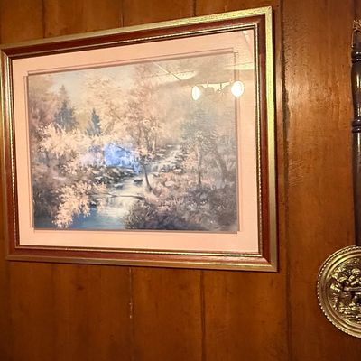 Framed landscape art print of a forest stream, flanked by a pair of vintage decorative brass warming pans with wooden handles.