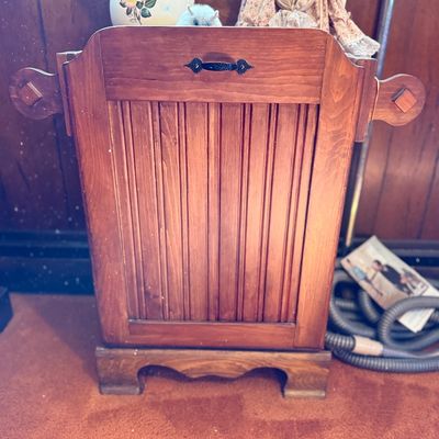 Vintage wooden tilt-out trash bin or hamper featuring beadboard paneling, decorative side handles, and a black metal pull, shown with...