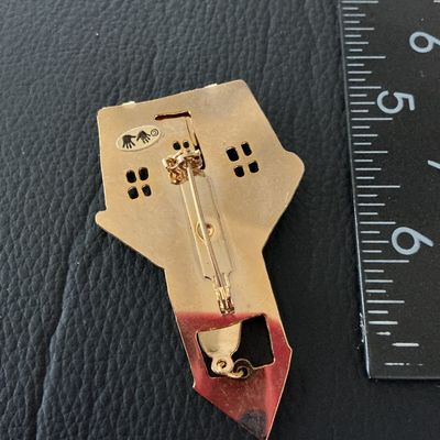 Sale Photo Thumbnail #151: A gold-tone and multicolored enamel brooch shaped like a red schoolhouse with a fence and a dangling green bell charm inside the belfry. Features a pin-back closure with standard rollover clasp. Marked on the back with a maker's mark showing two hands. Me