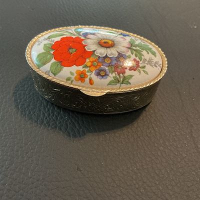 Sale Photo Thumbnail #219: Small vintage oval metal pill box featuring a colorful floral enamel top and etched scrollwork designs on the metal body and bottom. Opens via a hinged lid and includes a removable white plastic insert inside. Measures approximately 1.75 to 2 inches long.