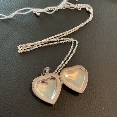 Sale Photo Thumbnail #225: A silver-tone rope chain necklace featuring a heart-shaped locket pendant. The front of the locket has a glittery enamel-style inlay bordered by clear rhinestones, and opens to reveal two picture slots. Includes an adjustable extender chain with a lobster