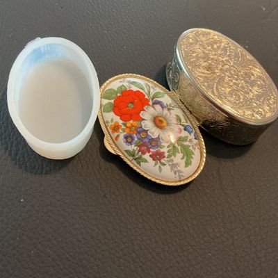 Sale Photo Thumbnail #221: Small vintage oval metal pill box featuring a colorful floral enamel top and etched scrollwork designs on the metal body and bottom. Opens via a hinged lid and includes a removable white plastic insert inside. Measures approximately 1.75 to 2 inches long.
